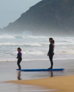 A woman and a girl at the rough sea wearing wetsuits standing on a surfboard on the sand