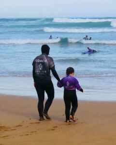 a woman and a girl walking hand in hand towards the ocean, wearing wetsuits